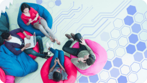 Four professionals relax on colorful beanbag chairs in a collaborative workspace, viewed from above as they work on laptops and mobile devices. Soft blue circuit lines and hexagon patterns overlay the floor, reinforcing a modern, tech-driven atmosphere built for connection and shared ideas.