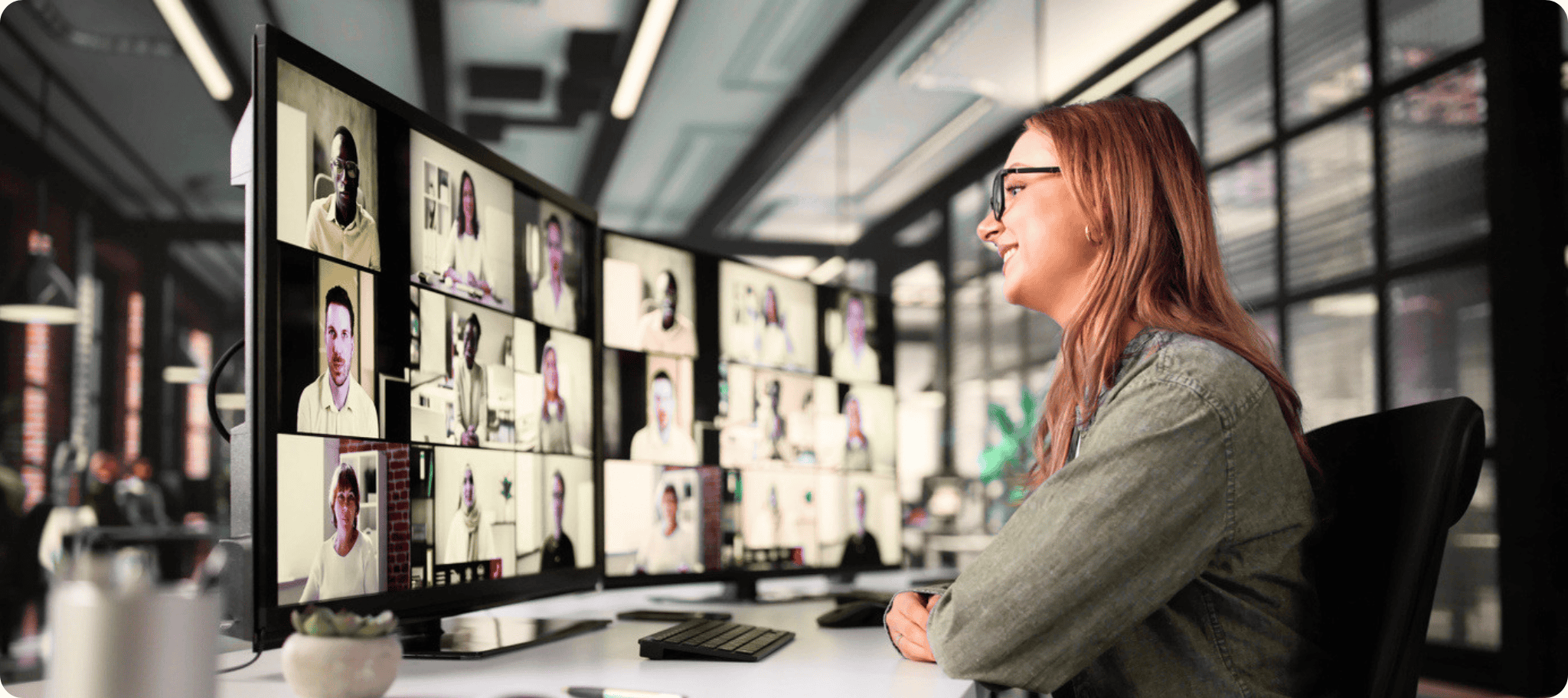 A woman sits at a desk in a modern office, smiling while participating in an AI Training video call displayed across multiple monitors showing a grid of people.