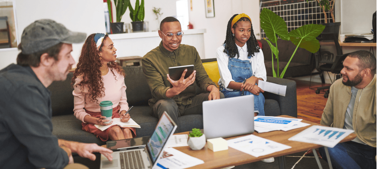 Five colleagues gather around a coffee table in a bright office, collaborating with laptops, notes, and printed charts as they share ideas in a relaxed team meeting.