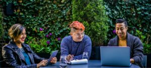 Group of three diverse staffing employees sitting outside, looking at LGBTQ+ career resources on laptop.