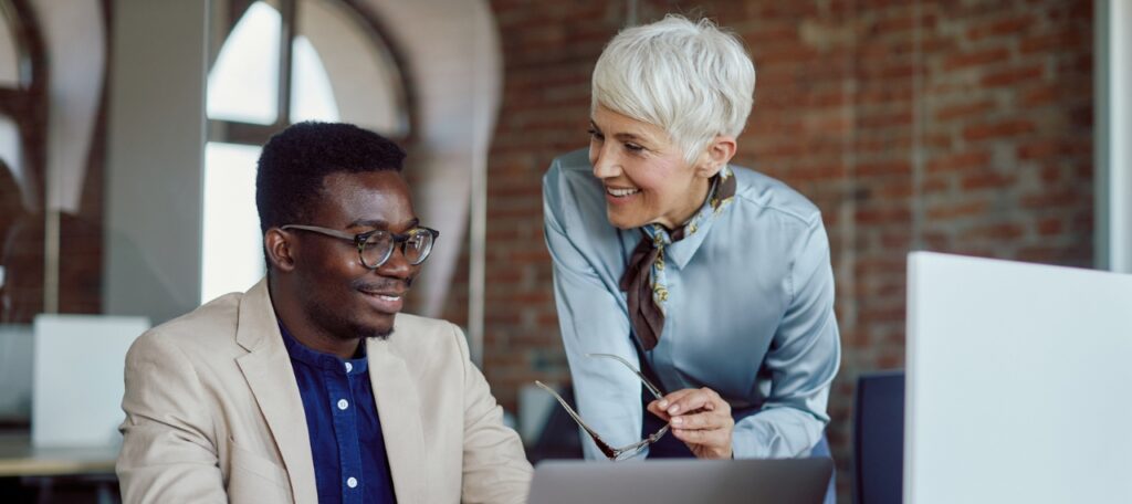 A smiling older woman with short white hair leans over to engage in conversation with a younger man wearing glasses and a beige blazer. They are in a modern office setting with exposed brick walls, natural light, and a laptop open in front of them. The image conveys a collaborative and inclusive work environment, reinforcing the theme of disability inclusion in recruitment.