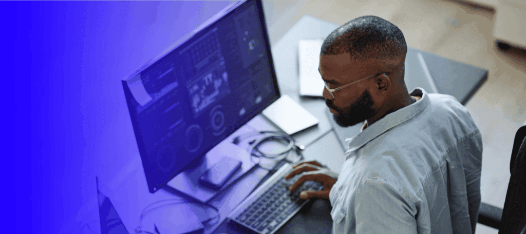 AI expert working at a desk, typing on a keyboard while viewing data dashboards on a widescreen monitor, with a blue gradient overlay on the left side of the image.