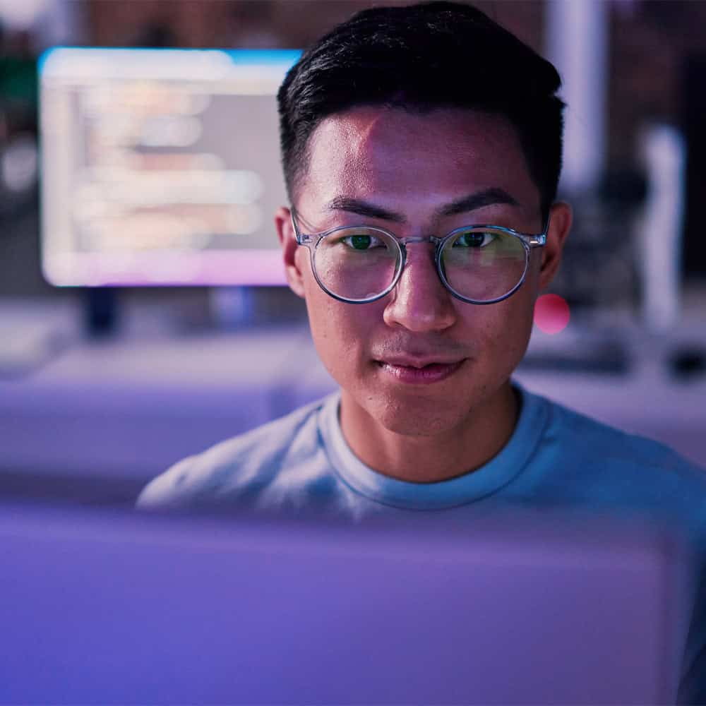 Close-up of a person wearing round glasses focused on a laptop screen while coding. A blurred monitor with lines of code appears in the background, suggesting a software development or engineering workspace. The person’s face is illuminated by the computer screen in a dimly lit environment.