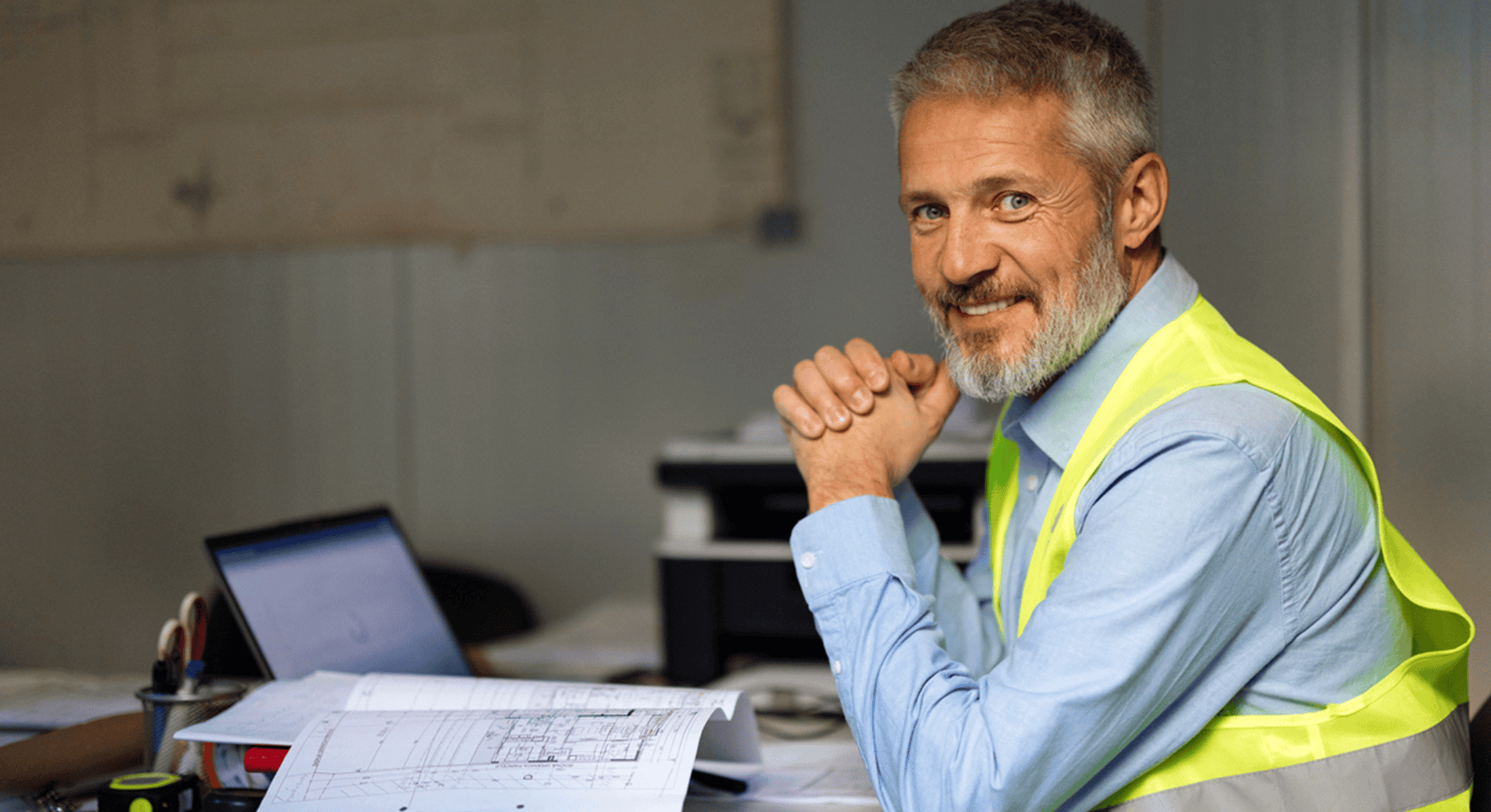 A smiling construction professional in a high-visibility safety vest sits at a desk reviewing architectural plans beside a laptop, creating a confident and approachable workplace scene that reflects hands-on project management, planning, and collaboration.