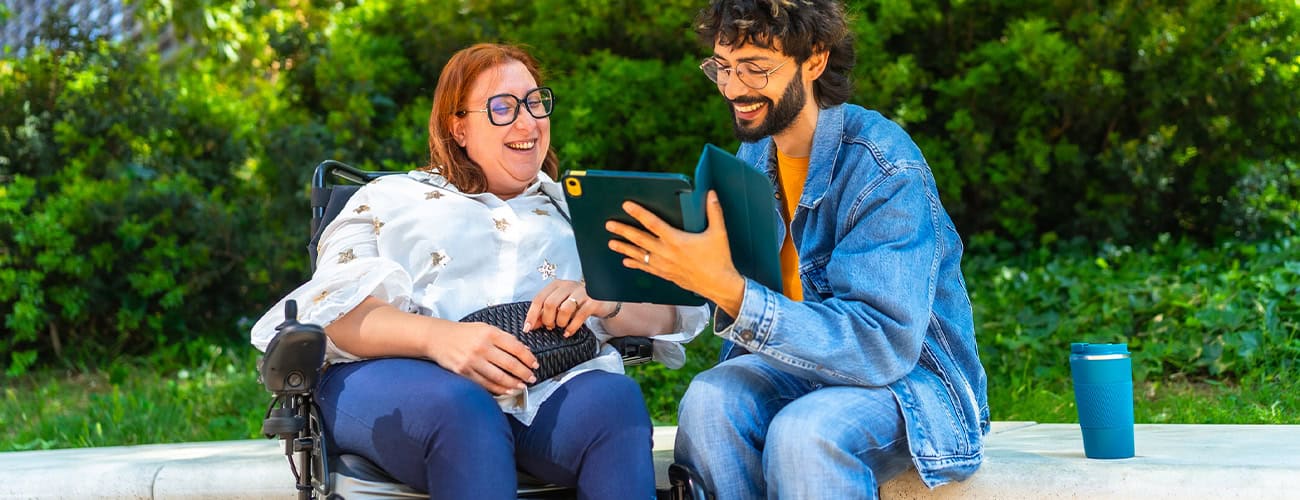Two professionals smiling outdoors while looking at a tablet, including one person using a wheelchair.