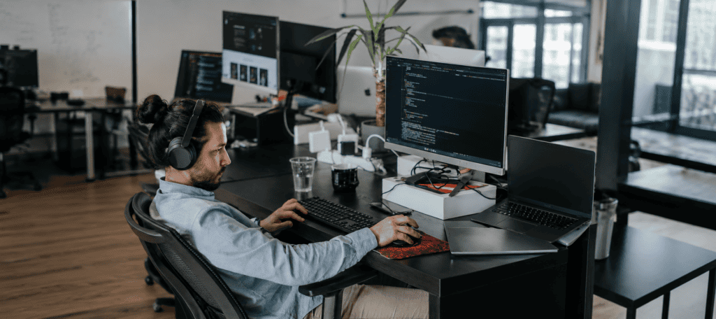 A focused software developer wearing headphones works across multiple monitors displaying code in a modern, open office, surrounded by laptops and technology that support a productive digital workspace.