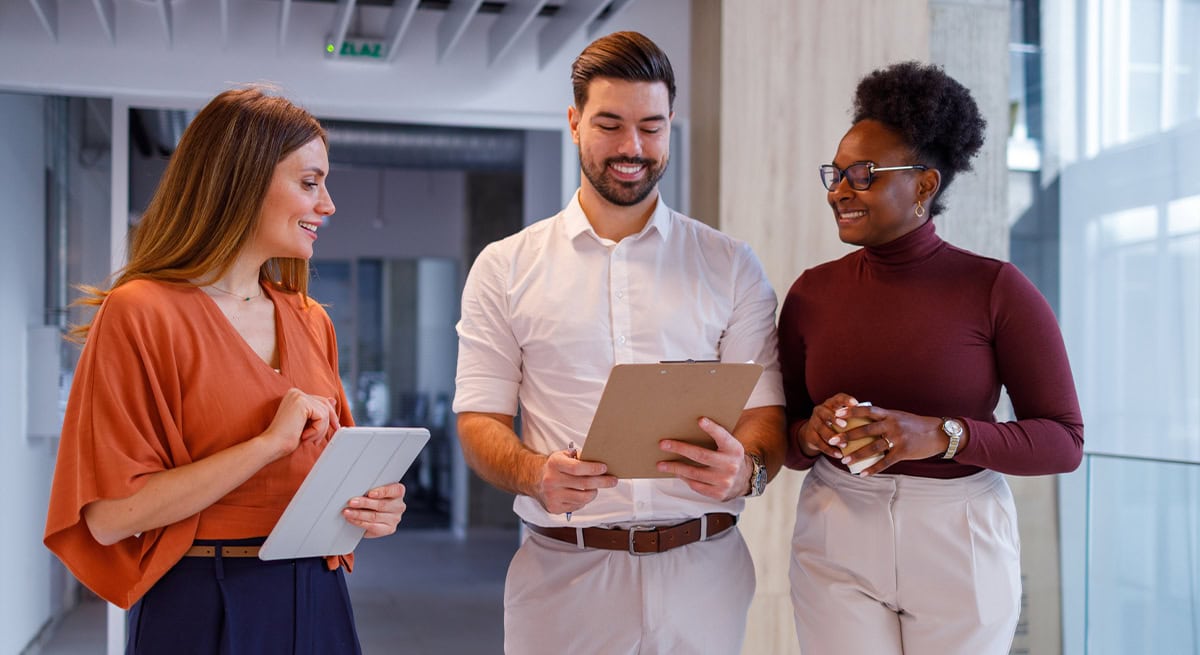Three business professionals collaborating in a modern office, reviewing documents on a clipboard and tablet, representing Administrative and Professional Contract Staffing.