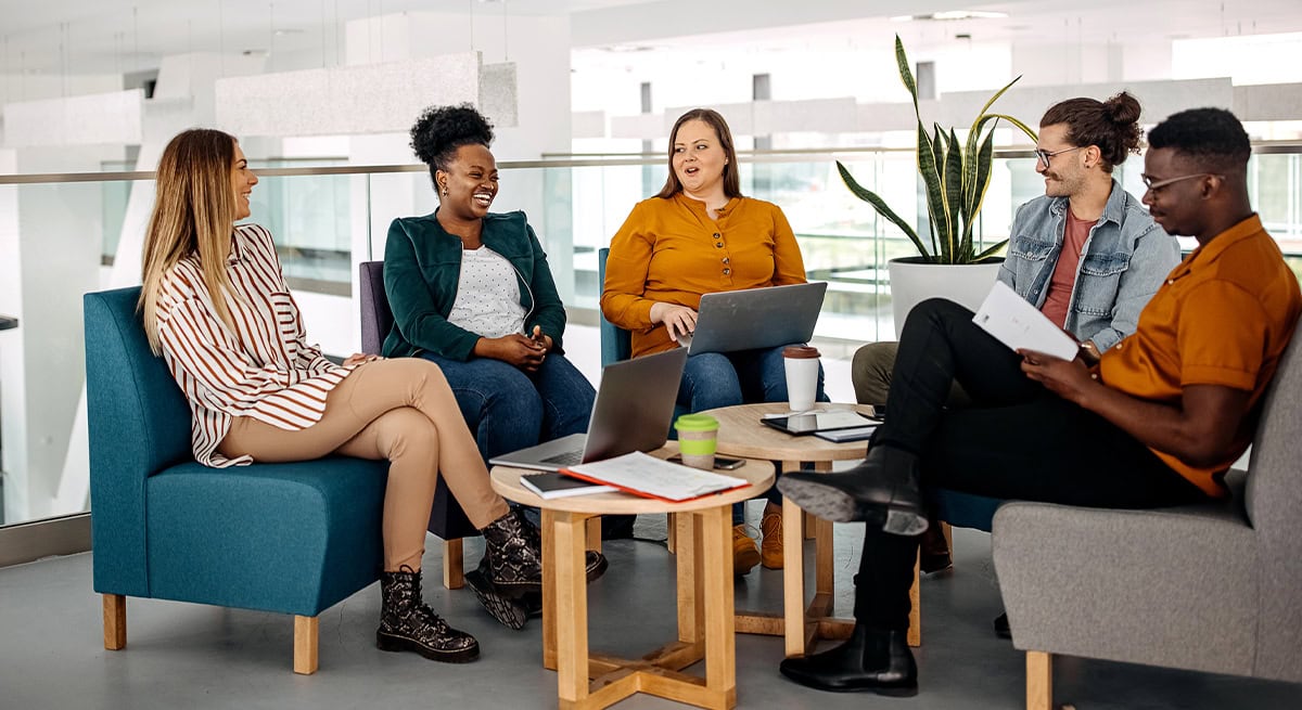 Five business professionals collaborating in a modern office lounge, discussing ideas and reviewing documents on laptops, representing Administrative Team Staffing.