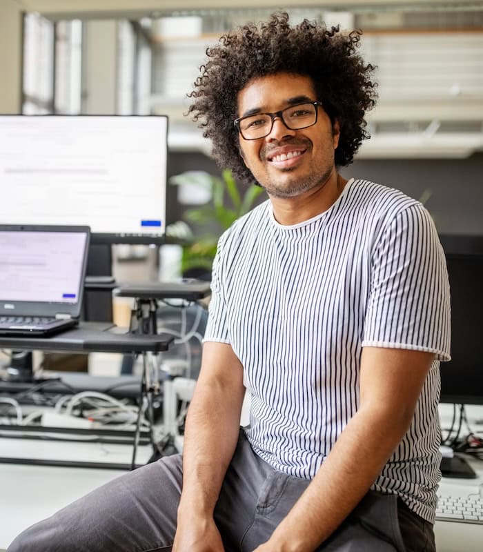 Smiling technology professional seated in a modern office with computer monitors and a laptop in the background, representing AI Skilled Talent.